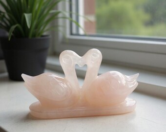 May include: A light pink swan ornament, featuring two swans in a heart shape. The translucent material of the figurine is visible. The swans are detailed with curved necks and wings, and sit on a rectangular base.