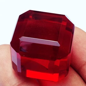 May include: A faceted, rectangular-cut, deep red gemstone. The gem has a high degree of clarity and brilliance, with multiple facets reflecting light. The gemstone is held in a hand, showcasing its size and color.