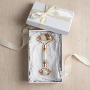 Puede incluir: Una caja de regalo blanca con una cinta color crema contiene un rodillo facial. El rodillo tiene un marco color cobre y un mango y rodillos de piedra blanca y marrón. La caja está forrada con papel de seda blanco.