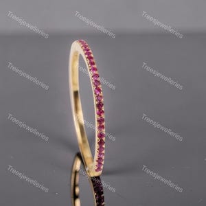 May include: A gold band ring with a row of small, vibrant pink gemstones set along the top. The ring is displayed on a reflective surface, showcasing its polished finish and the sparkle of the gemstones. The ring is a piece of jewelry.