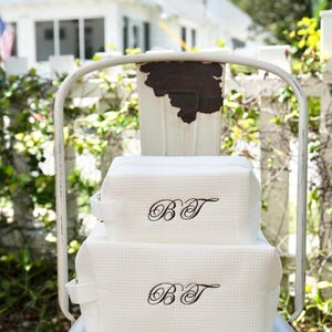 Peut inclure: Deux trousses de toilette blanches et texturées, avec les initiales brodées en noir "BT", sont empilées sur une chaise en métal blanc. Les sacs ont de petites poignées et une forme rectangulaire. En arrière-plan, une clôture et une maison blanches.