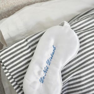 May include: A white satin sleep mask with the embroidered phrase "Do Not Disturb" in blue script. The mask rests on a striped gray and white pillowcase, with a white pillow and a neutral-toned surface in the background.