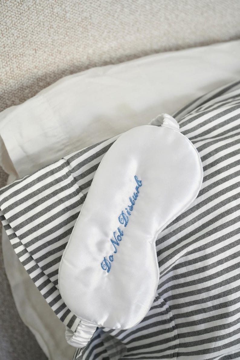 Op de afbeelding: Een witte satijnen slaapmasker met de geborduurde tekst "Do Not Disturb" in blauw. Het masker ligt op een gestreepte grijs-witte kussensloop, met een wit kussen en een neutrale achtergrond.