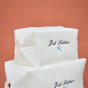 May include: Two white, textured travel bags stacked on top of each other. The bags have a rectangular shape with a zipper closure and a handle. The words "Jet Setter" are embroidered in black script with a blue airplane.