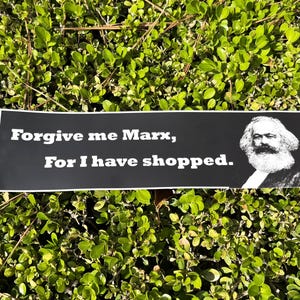 May include: A black bumper sticker with white text that reads "Forgive me Marx, For I have shopped." A black and white portrait of Karl Marx is on the right side of the sticker. The sticker is set against a background of green foliage.