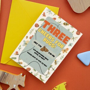 May include: An invitation with the text "THREE CHEERS FOR BUGS!" on a light blue background, surrounded by illustrations of insects. A yellow envelope and pen are next to the invitation, along with wooden toys and a blue triangle on an orange surface.