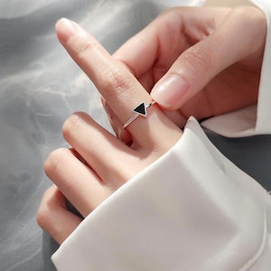 Peut inclure: Une bague en argent avec une pierre noire en forme de triangle. La bague est portée au doigt, avec une chemise blanche en arrière-plan. Le design de la bague est minimaliste et géométrique, adapté au quotidien.