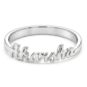 Puede incluir: Un anillo plateado con el nombre "Akarsha" en escritura cursiva. El anillo es una banda simple con el nombre como punto focal, sobre un fondo blanco.