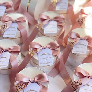 May include: Ivory round gift boxes with gold trim, tied with pink satin bows and ribbons. Each box has a tag with gold lettering and a message. The boxes are arranged on a light surface.
