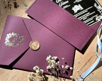 Pode incluir: Conjunto de convite de casamento cor de ameixa com um selo de cera dourada e enfeites florais. O convite apresenta um cartão preto com texto branco. Um fio de pérolas e uma fita branca contribuem para a apresentação elegante.