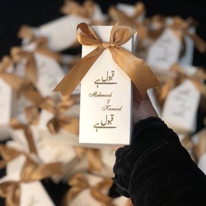 May include: A white gift box tied with a gold ribbon, with gold script text. The box is held in front of a blurred background of similar boxes. The text on the box reads "Mahmood & Kanwal".