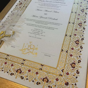 以下が含まれることがあります： 金とバーガンディの花柄の縁取りが施された、額入りの結婚証明書。 英語とアラビア語の優雅なカリグラフィーで、新郎新婦の名前が記されています。 装飾用の羽と金の装飾が、フォーマルな美しさを添えています。