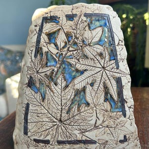 May include: A cream-coloured ceramic sculpture with a flat, trapezoidal shape. The surface features intricate, embossed leaf patterns and dark brown outlines, with patches of glossy blue glaze filling the negative spaces between the foliage.