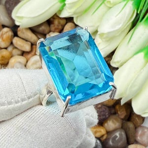 Puede incluir: Un anillo de plata con una gran piedra preciosa de color azul cielo, rectangular y facetada, sujeta por cuatro garras. La piedra muestra un tono azul vibrante y translúcido con bordes nítidos y un acabado pulido sobre una banda de plata sencilla.