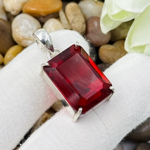 May include: A rectangular, faceted deep red gemstone pendant set in a silver-toned metal frame with a simple bail at the top. The stone features sharp, clean edges and a polished surface, reflecting light across its vibrant, saturated crimson color.