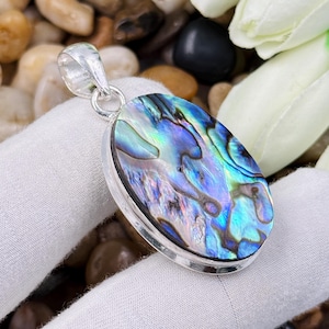 Può includere: Un ciondolo ovale con un inserto in conchiglia di abalone lucidata, caratterizzato da vortici iridescenti blu, verdi e viola. La conchiglia è incastonata in una cornice di metallo color argento liscio e presenta un piccolo occhiello circolare.