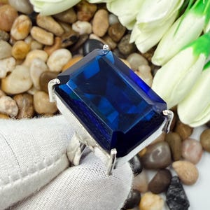 Puede incluir: Un anillo con una gran piedra preciosa rectangular facetada de color azul profundo, montada en un engaste de metal plateado con garras. La piedra muestra un tono azul vibrante y saturado con bordes limpios y una superficie pulida.