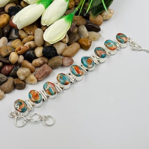 Könnte beinhalten: Ein silbernes Gliederarmband mit neun ovalen Steinen in einem marmorierten Muster aus Türkis-, Orange- und Kupferfarben. Das Armband verfügt über einen Knebelverschluss und eine kurze Verlängerungskette mit kreisförmigen Gliedern am Ende.
