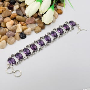 May include: A silver link bracelet featuring eight oval-cut purple amethyst gemstones. Each stone is set in a decorative silver frame with small beaded accents. The bracelet includes a toggle clasp closure with a circular ring and a T-bar.