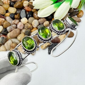 Può includere: Un bracciale a fascia in argento con quattro pietre preziose di peridoto verde lime, tagliate a sfaccettature ovali. Ogni pietra è incastonata in una montatura in metallo intrecciato. Il bracciale ha un design aperto con finitura in argento lucido.