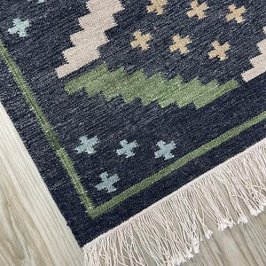 May include: A close-up of a navy blue and beige patterned rug with a green border and white fringe. The rug features a geometric design with cross-like shapes in beige and light blue. The rug is made of woven fabric.
