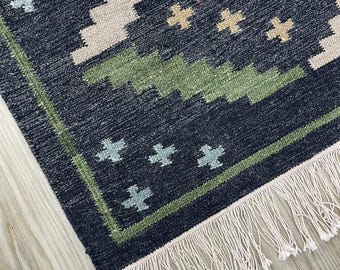 Puede incluir: Primer plano de una alfombra estampada en azul marino y beige con borde verde y flecos blancos. La alfombra presenta un diseño geométrico con formas de cruz en beige y azul claro. La alfombra está hecha de tela tejida.