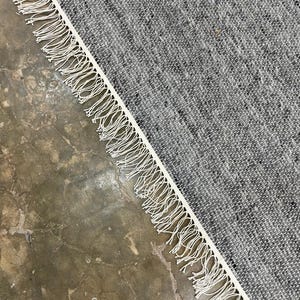 May include: A grey and white woven rug with a fringed border. The rug has a textured appearance and is angled across a concrete surface. The fringe is a bright white colour.