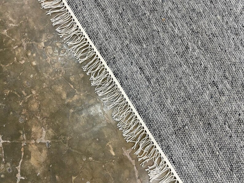 May include: A grey and white woven rug with a fringed border. The rug has a textured appearance and is angled across a concrete surface. The fringe is a bright white colour.