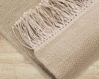 May include: Beige woven rug with a textured surface and a fringe border. The rug is folded over, revealing the underside. The neutral colour and simple design make it suitable for various interior styles.