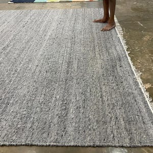 May include: A large, rectangular grey rug with a textured surface. The rug has a fringe border and is made of woven material. The rug is displayed on a concrete floor.