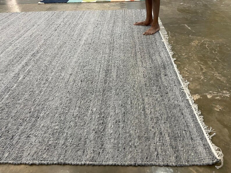 May include: A large, rectangular grey rug with a textured surface. The rug has a fringe border and is made of woven material. The rug is displayed on a concrete floor.