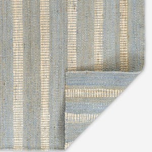 Può includere: Un tappeto intrecciato caratterizzato da un motivo a righe verticali in tonalità alternate di azzurro e beige naturale. Il tappeto presenta una trama grossolana con un angolo ripiegato che rivela il retro del tessuto.