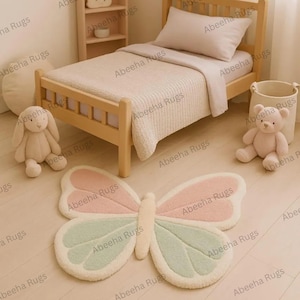 Könnte beinhalten: Ein schmetterlingsförmiger Teppich in Pastellrosa und Mintgrün liegt auf einem hellen Holzboden. Ein helles Holzbett mit weißer Bettdecke und Kissen befindet sich in der Nähe. Ein Stoffhase und ein Teddybär sind ebenfalls im Raum.