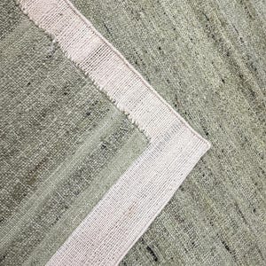 Puede incluir: Primer plano de una alfombra tejida en verde claro y beige. La alfombra tiene una textura visible con un patrón de tejido. Un borde beige enmarca el borde, agregando un elemento de contraste al diseño. La textura de la alfombra es claramente visible.