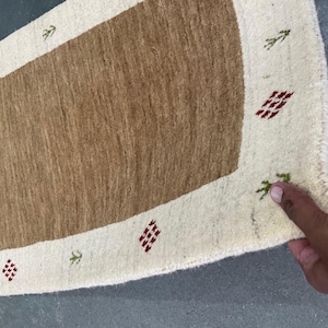 May include: A rectangular rug with a central brown section and an off-white border. The border features small red diamond patterns and green leaf-like designs. The rug is made of a woven material.