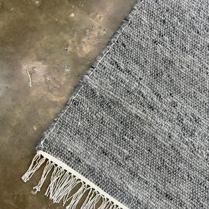 May include: A corner of a grey woven rug with a white border and fringe. The rug has a textured appearance with flecks of darker grey. The white fringe hangs along the bottom edge.