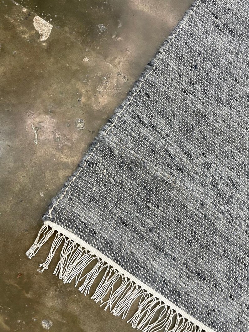 May include: A corner of a grey woven rug with a white border and fringe. The rug has a textured appearance with flecks of darker grey. The white fringe hangs along the bottom edge.