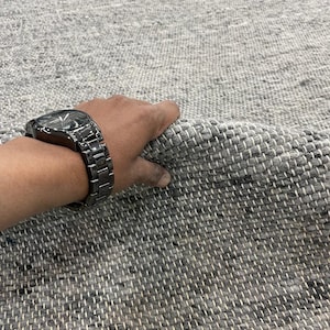 May include: A grey woven rug with a textured surface. The rug is made of a light and dark grey material, with a woven pattern. The rug is being held up by a hand wearing a black watch.