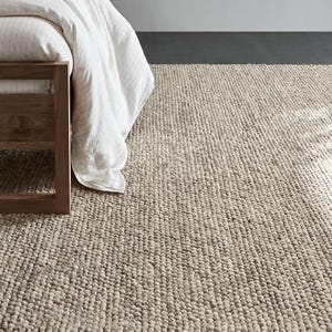 May include: A neutral-toned, textured area rug with a woven design. The rug is made of a thick, durable material and has a soft, inviting appearance. A wooden bed frame and white bedding are partially visible.