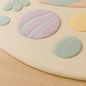 Puede incluir: Primer plano de una alfombra redonda de color crema con formas en colores pastel. La alfombra presenta c&iacute;rculos y dise&ntilde;os en forma de p&eacute;talos en tonos rosa, amarillo, verde y azul. La alfombra est&aacute; colocada sobre un suelo de madera clara.