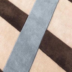 Op de afbeelding: Een geweven vloerkleed met een geometrisch patroon van elkaar kruisende diagonale strepen. Het ontwerp bestaat uit brede banen in donkerbruin, lichtbeige en gedempt leiblauw, wat een gelaagd, kruisend effect over het oppervlak creëert.
