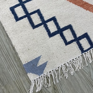 Könnte beinhalten: Cremefarbener gewebter Teppich mit einem geometrischen Muster in Marineblau, Rost und Hellblau. Der Teppich hat Rauten- und Pfeilformen mit Fransen an einer Kante. Der Teppich ist wahrscheinlich aus Naturfasern gefertigt.