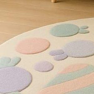 Puede incluir: Una alfombra redonda de color crema con caramelos y formas de colores pastel. La alfombra presenta dise&ntilde;os en azul claro, rosa, morado y amarillo. La alfombra est&aacute; hecha de un material suave y afelpado.