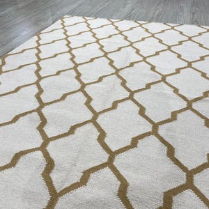 Könnte beinhalten: Ein weißer Teppich mit einem sich wiederholenden geometrischen Muster in einem warmen Braunton. Der Teppich besteht aus einem gewebten Material und hat eine strukturierte Oberfläche. Das Muster besteht aus ineinandergreifenden Formen.