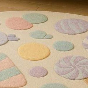 Puede incluir: Una alfombra semicircular de color crema con dise&ntilde;os de caramelos pastel. La alfombra presenta varias formas y colores, incluyendo rosa, morado, amarillo y verde, que se asemejan a piruletas y caramelos. La alfombra est&aacute; dise&ntilde;ada para la habitaci&oacute;n de un ni&ntilde;o o sala de juegos.