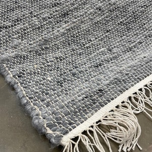 May include: Close-up of a handwoven rug with a textured surface. The rug features a mix of grey and white threads, creating a speckled pattern. The edge has a white border with long, off-white fringe.