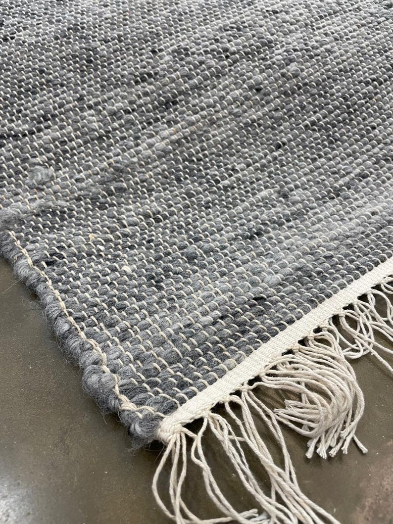 May include: Close-up of a handwoven rug with a textured surface. The rug features a mix of grey and white threads, creating a speckled pattern. The edge has a white border with long, off-white fringe.