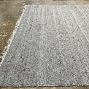 May include: A large, rectangular grey rug with a textured surface. The rug has a fringe detail along one edge and is made of woven material. The colour is a solid grey with subtle variations in tone.
