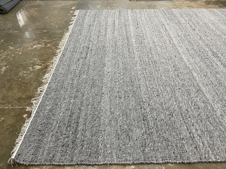 May include: A large, rectangular grey rug with a textured surface. The rug has a fringe detail along one edge and is made of woven material. The colour is a solid grey with subtle variations in tone.