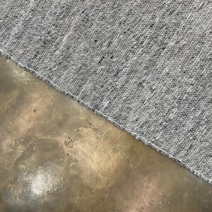 May include: Close-up of a grey textile with a textured weave, featuring flecks of black and white. The fabric's edge is finished with a white seam. The textile is set against a mottled, brown surface.
